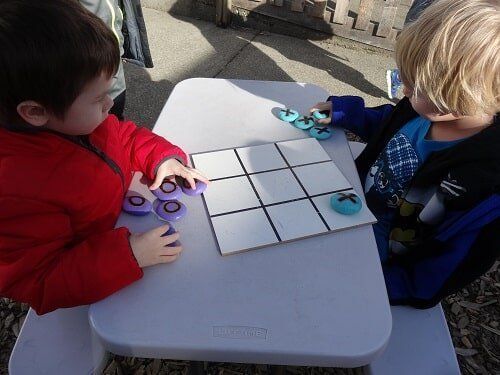Two young children playing tic-tac-toe at a white table outdoors, one in red jacket, the other in a blue jacket