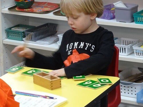 Young boy, seated at table, playing with number tiles and wooden box in a classroom setting.