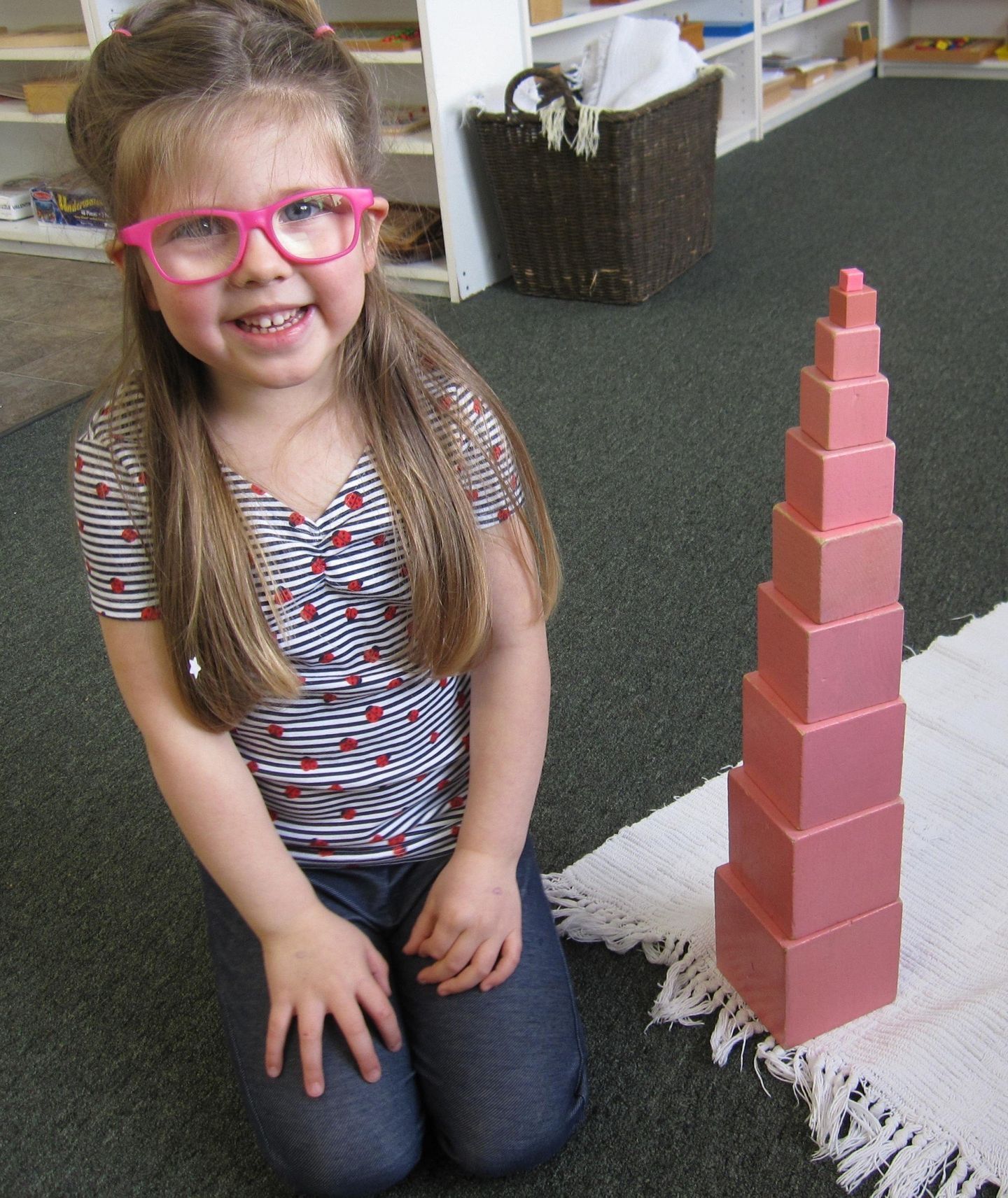 Young girl kneeling, smiling, next to a tower of pink blocks on a rug