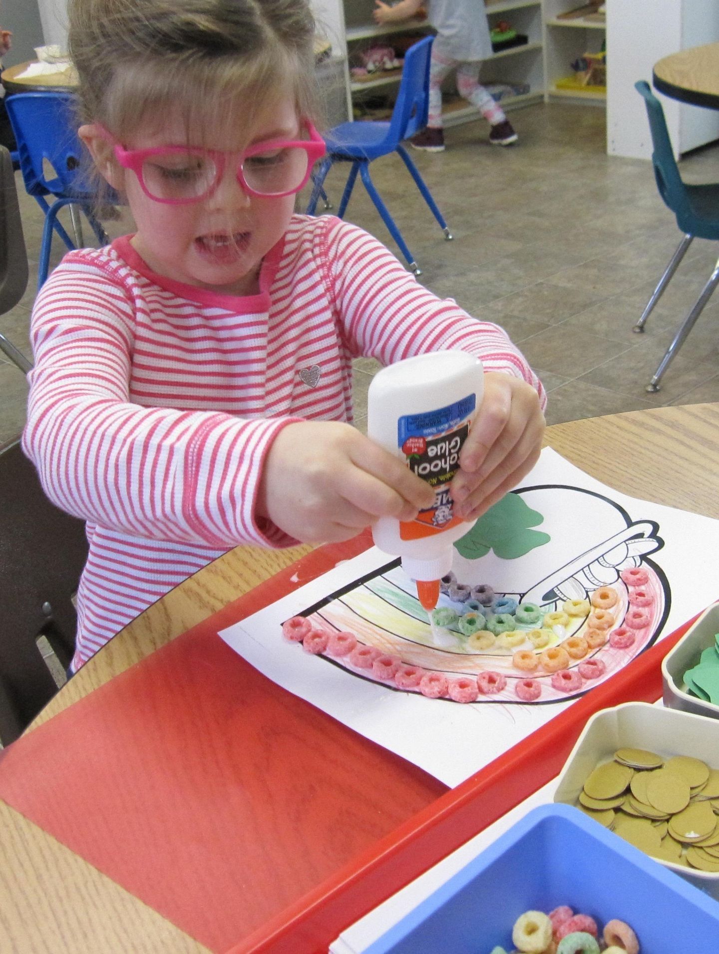 Girl with pink glasses glues cereal pieces onto a rainbow art project at a table