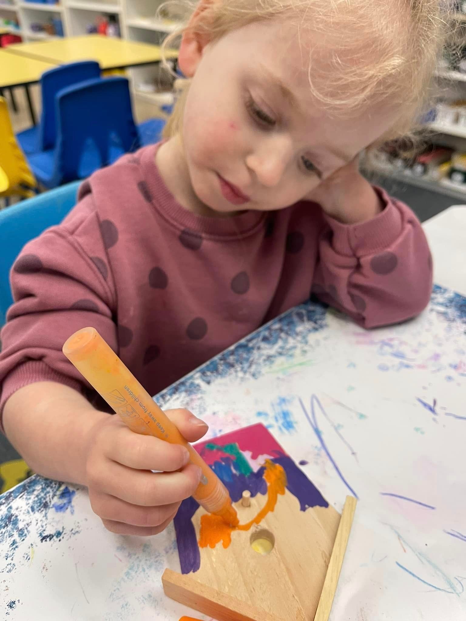 A young child colors a wooden birdhouse with an orange marker at a table