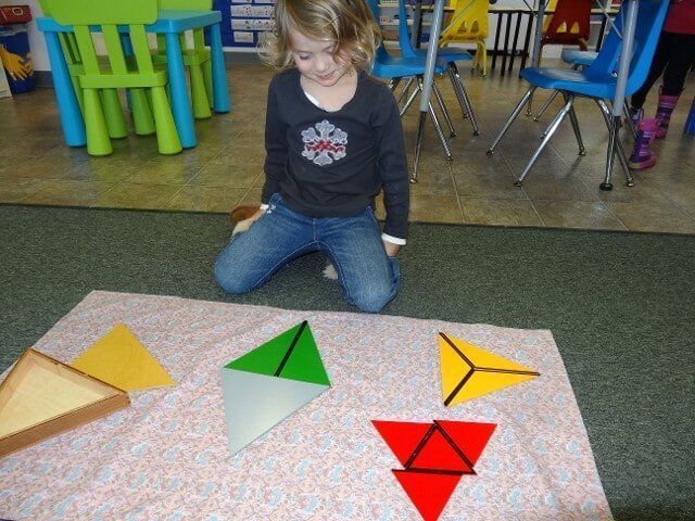 A child kneels, observing colorful geometric shapes on a patterned blanket in a classroom