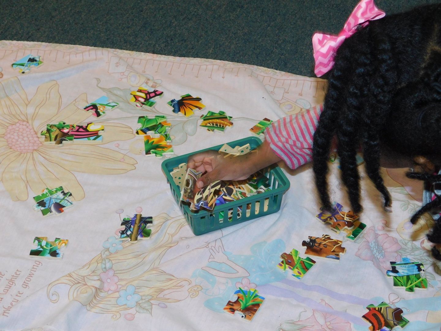 Child with braids and a pink bow, sorting puzzle pieces on a floral quilt, holding a green basket
