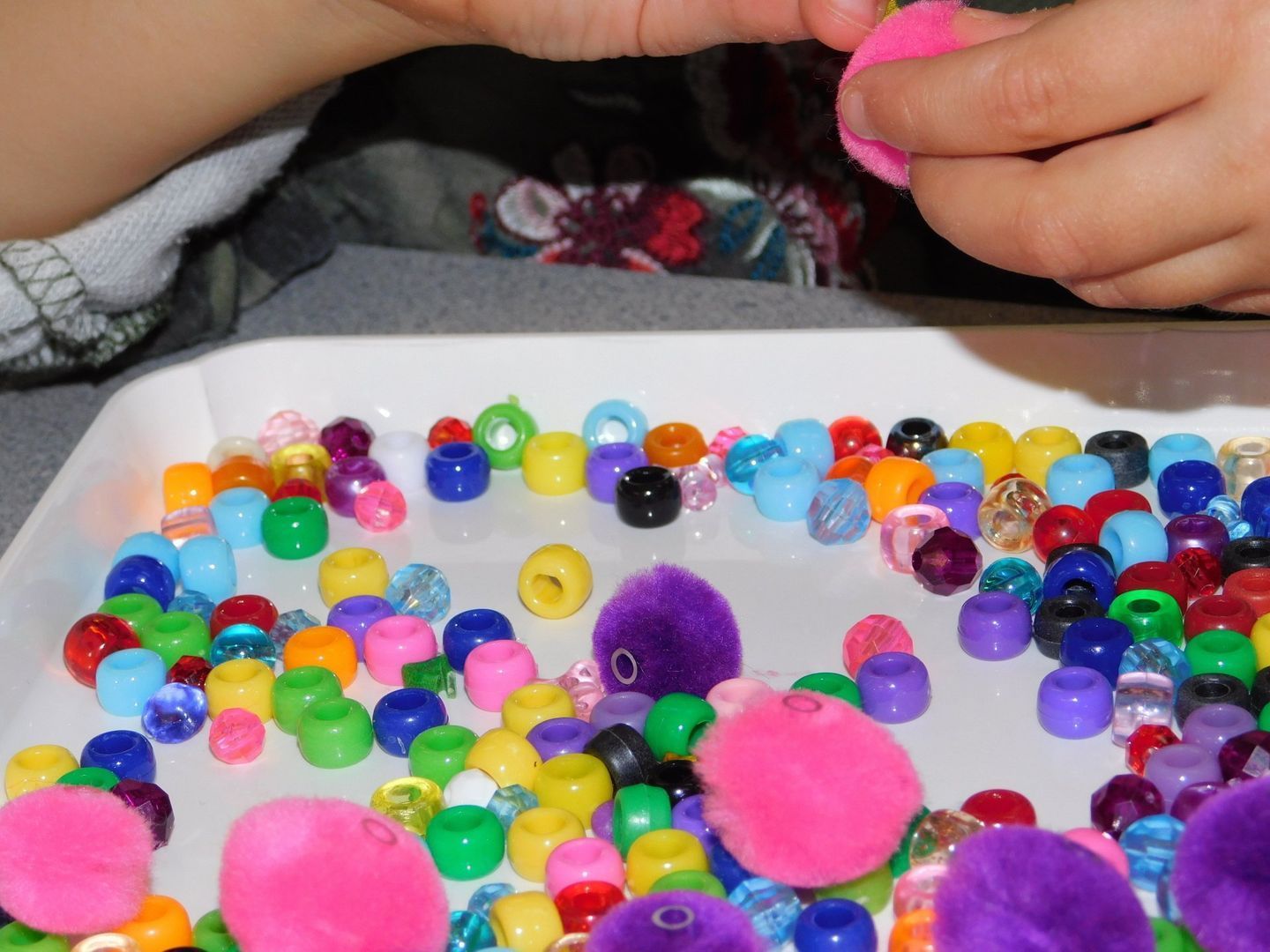 Child's hands stringing colorful beads onto a pink pompom in a tray of beads