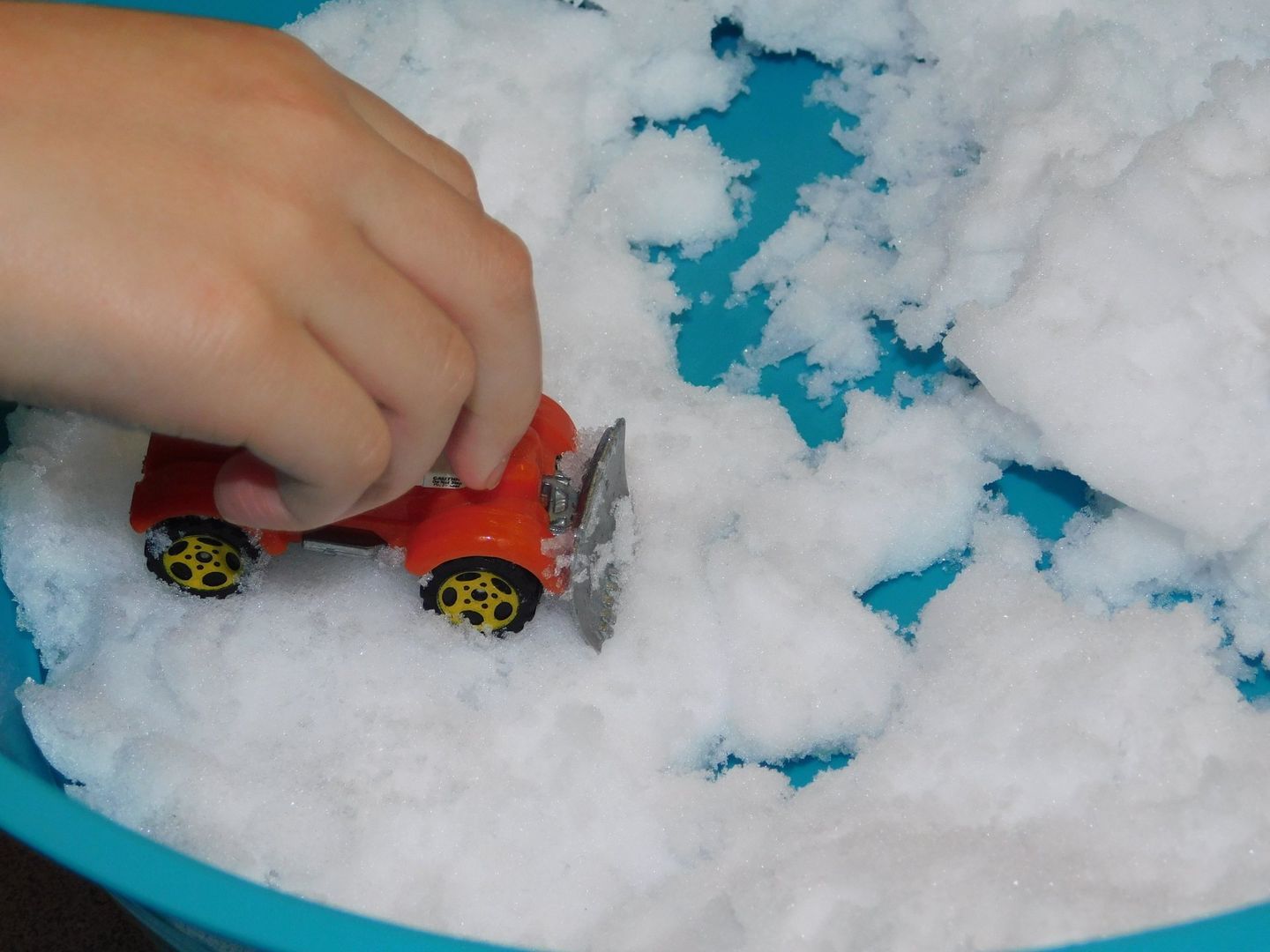 Child's hand uses a toy truck with a snowplow to clear fake snow in a blue bowl