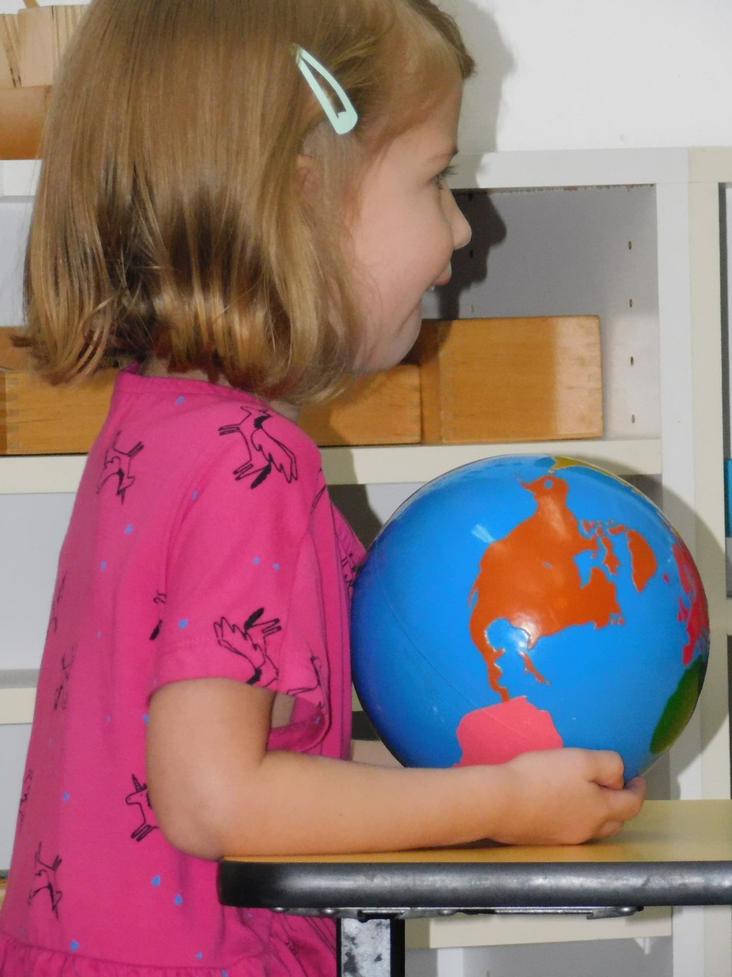 Girl in pink dress holding a blue globe, smiling in a classroom setting