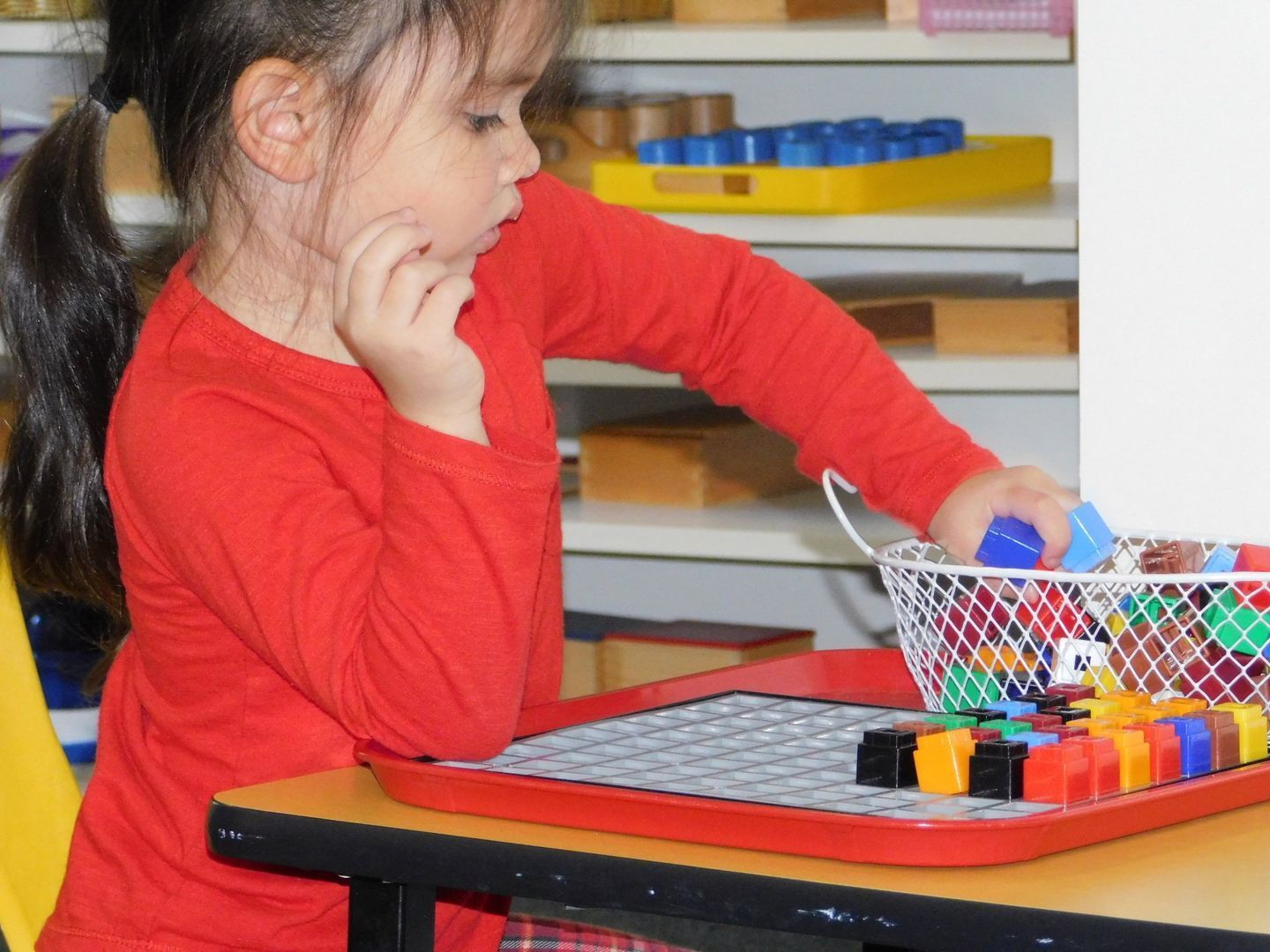 A young girl in red shirt sorting colorful blocks at a table in a classroom