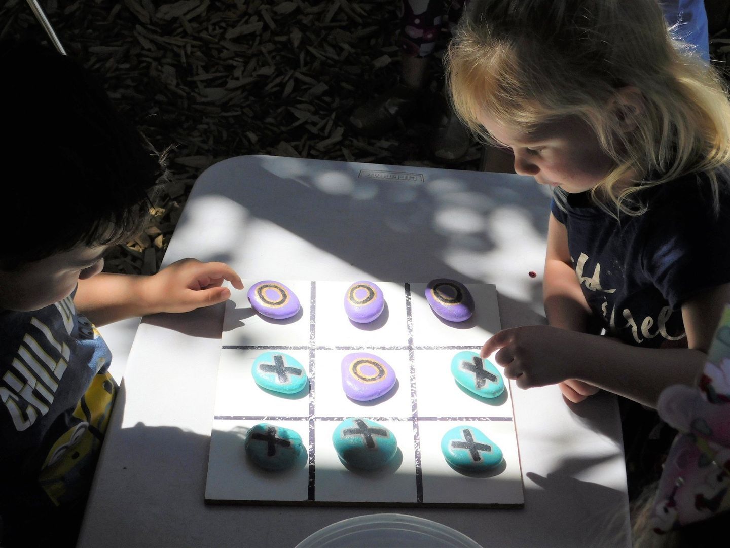 Two children playing tic-tac-toe with painted rocks on a white table outdoors