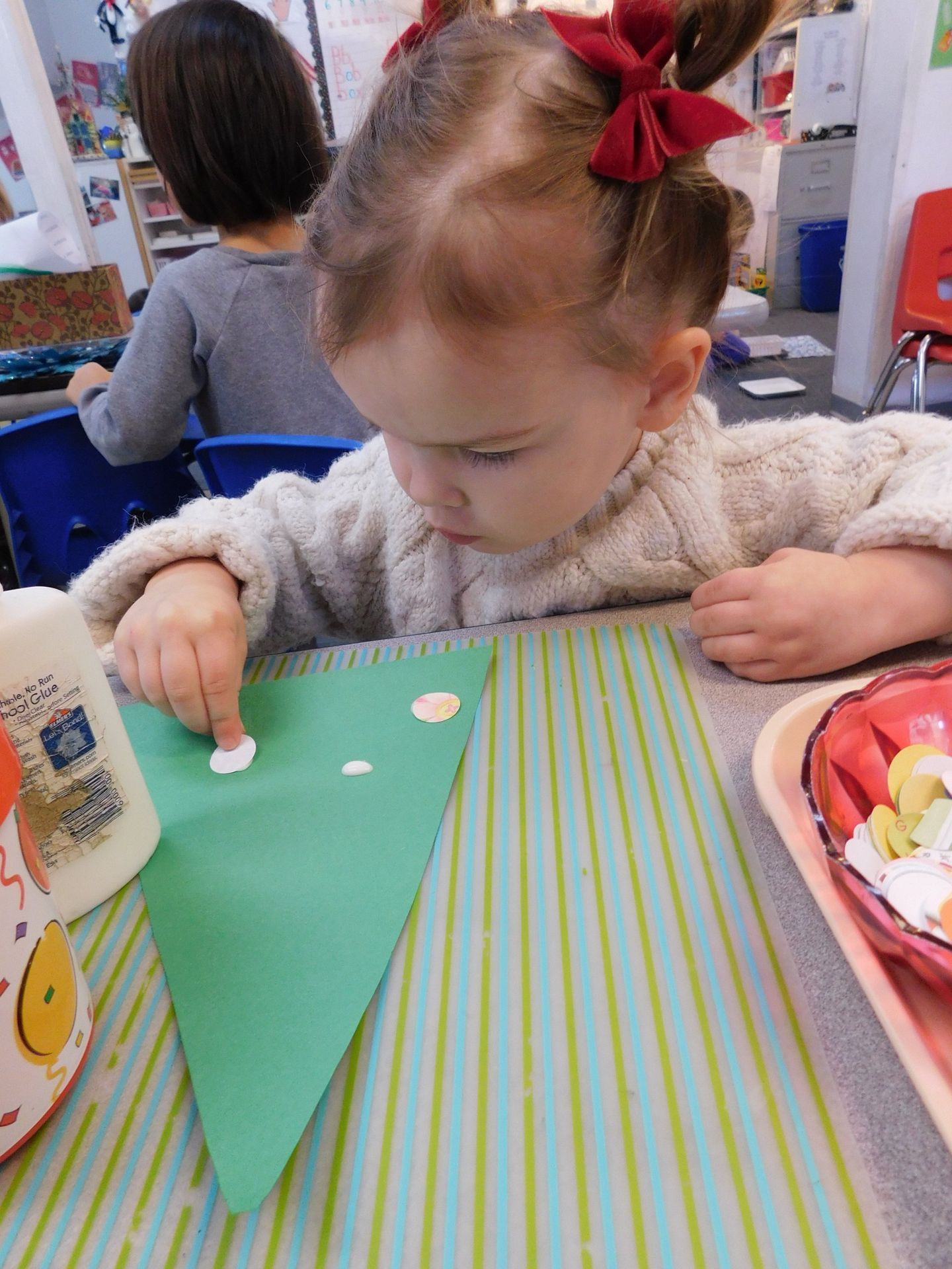 Girl with red bows putting white dots on green paper, at a table with other craft items