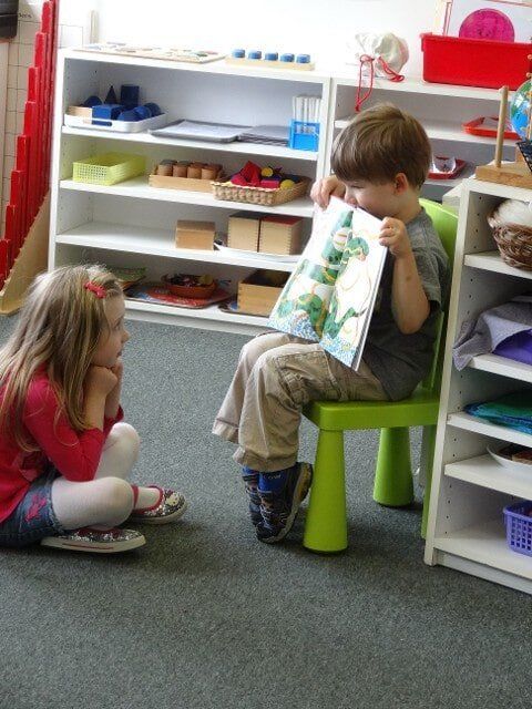 Girl sits, watching a boy read a book in a classroom