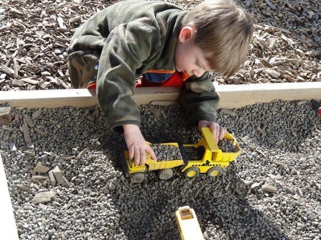 Young child playing with a yellow dump truck in a gravel-filled sandbox