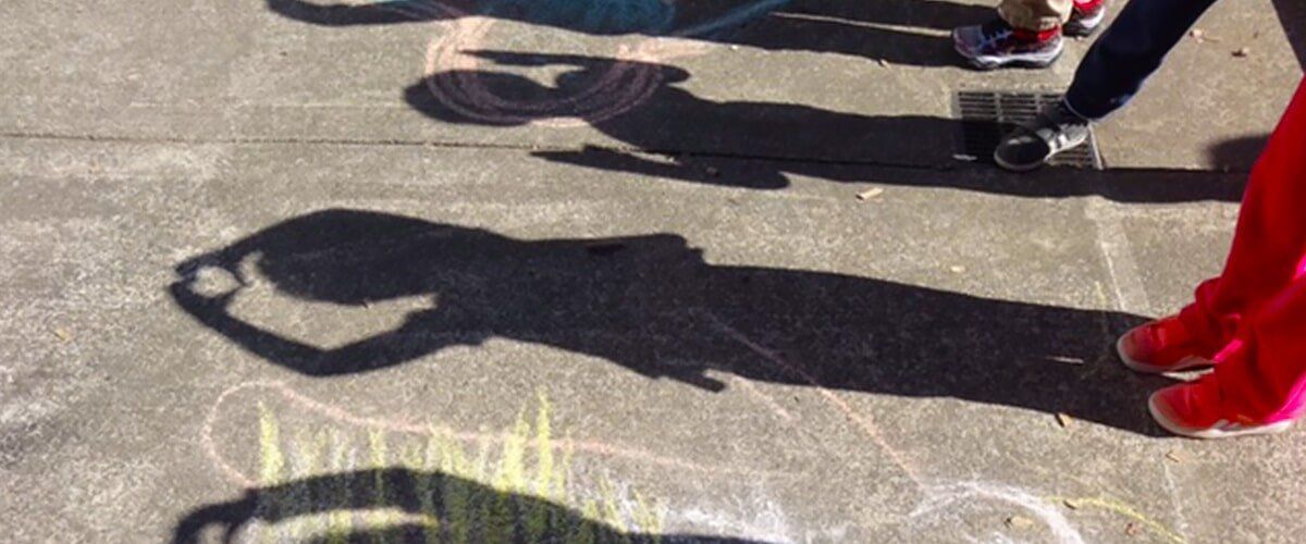 Shadows of people on a sidewalk, with colorful chalk drawings in the background