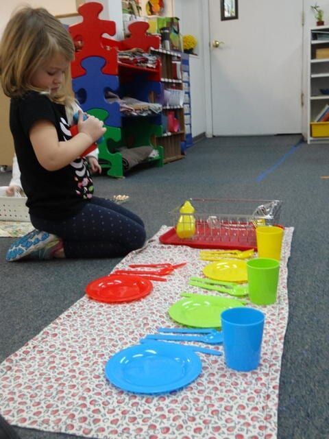 Girl setting colorful plates and cups on a patterned mat on the floor of a room with shelves and toys
