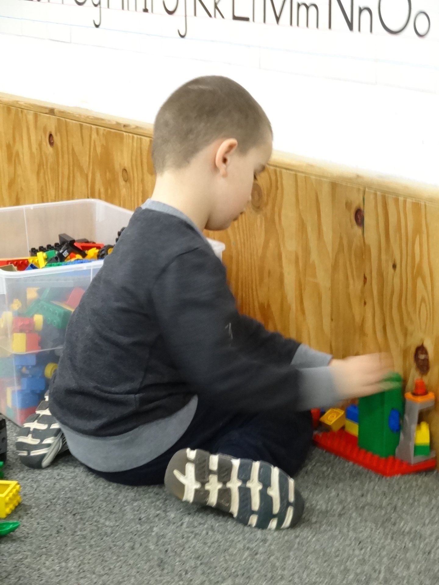 A young boy sitting on a carpet playing with colorful building blocks near a wooden wall