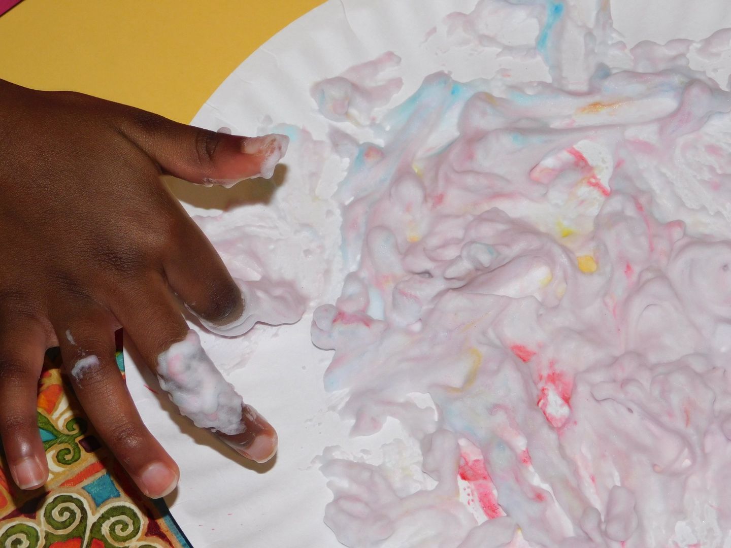A child's hand covered in white, colorful shaving cream on a paper plate