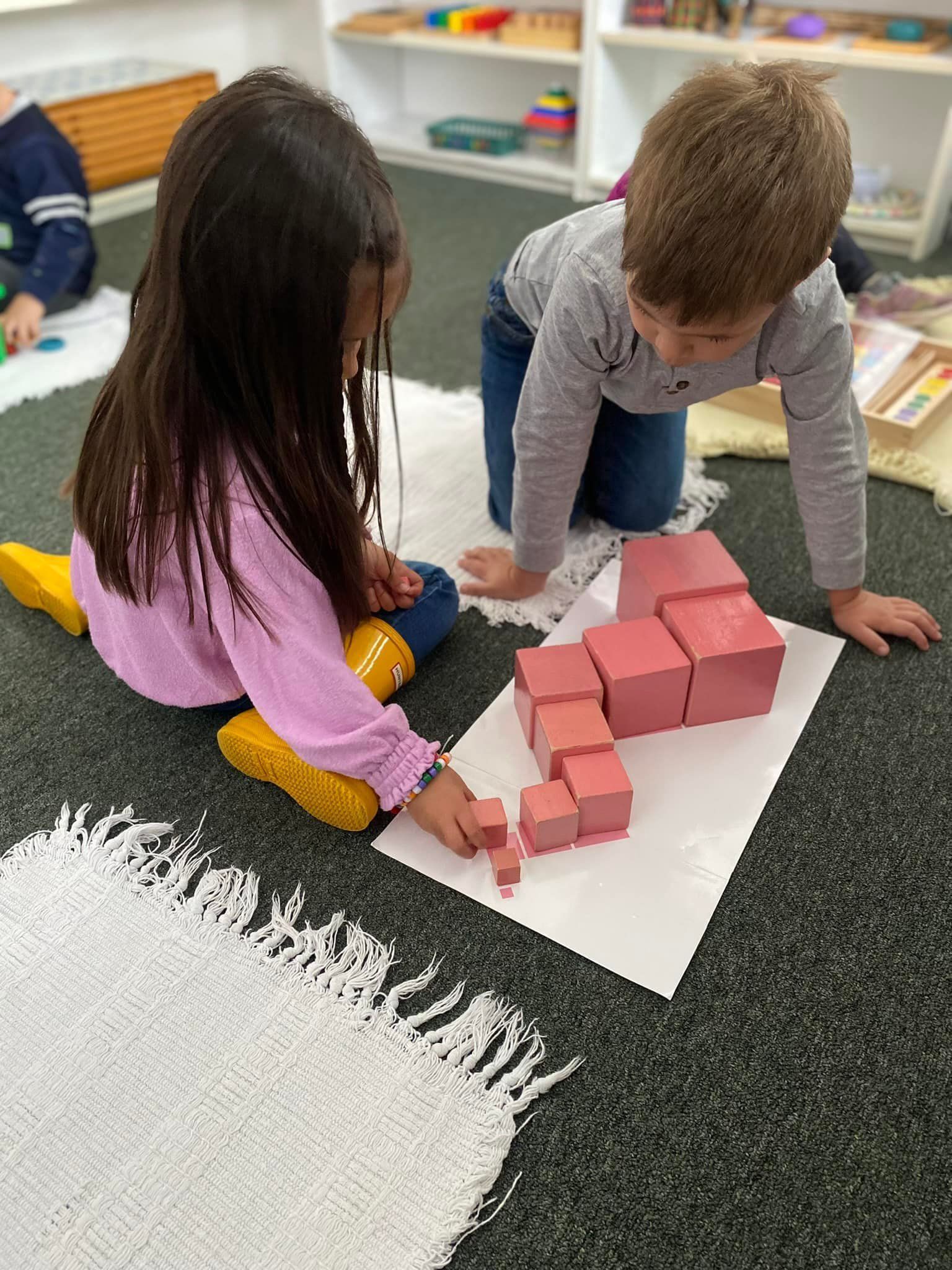 Two children building with pink blocks on a white paper, seated on a rug