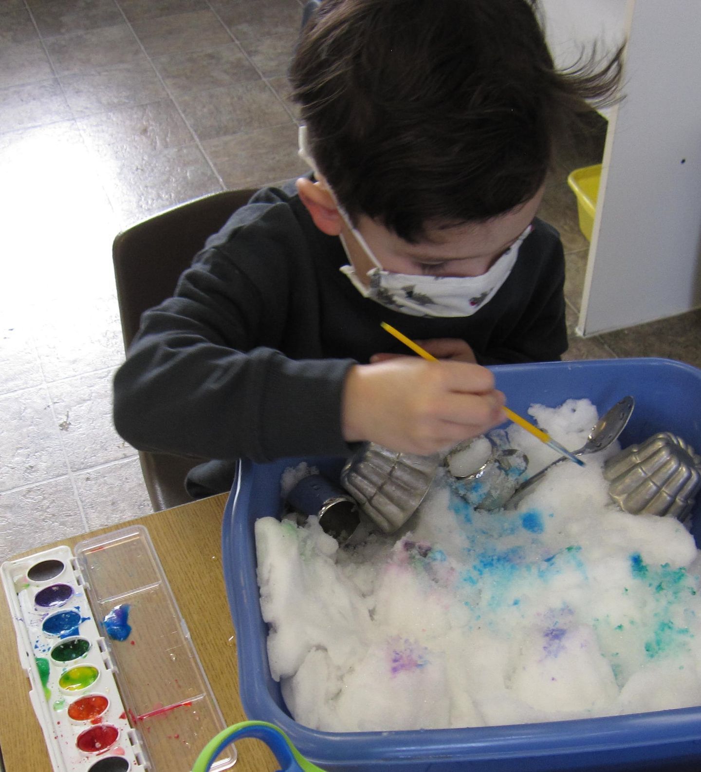 A child wearing a mask paints cotton balls with watercolors in a blue bin at a table