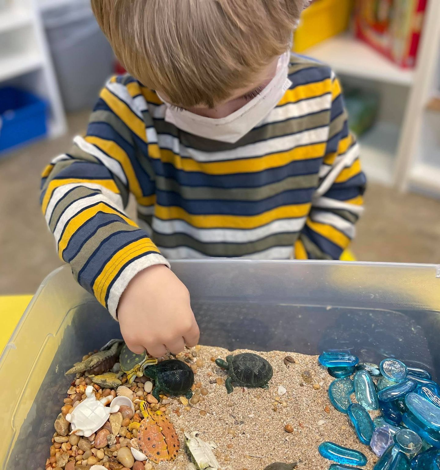 A young child wearing a mask plays with sand, pebbles, and turtles in a bin