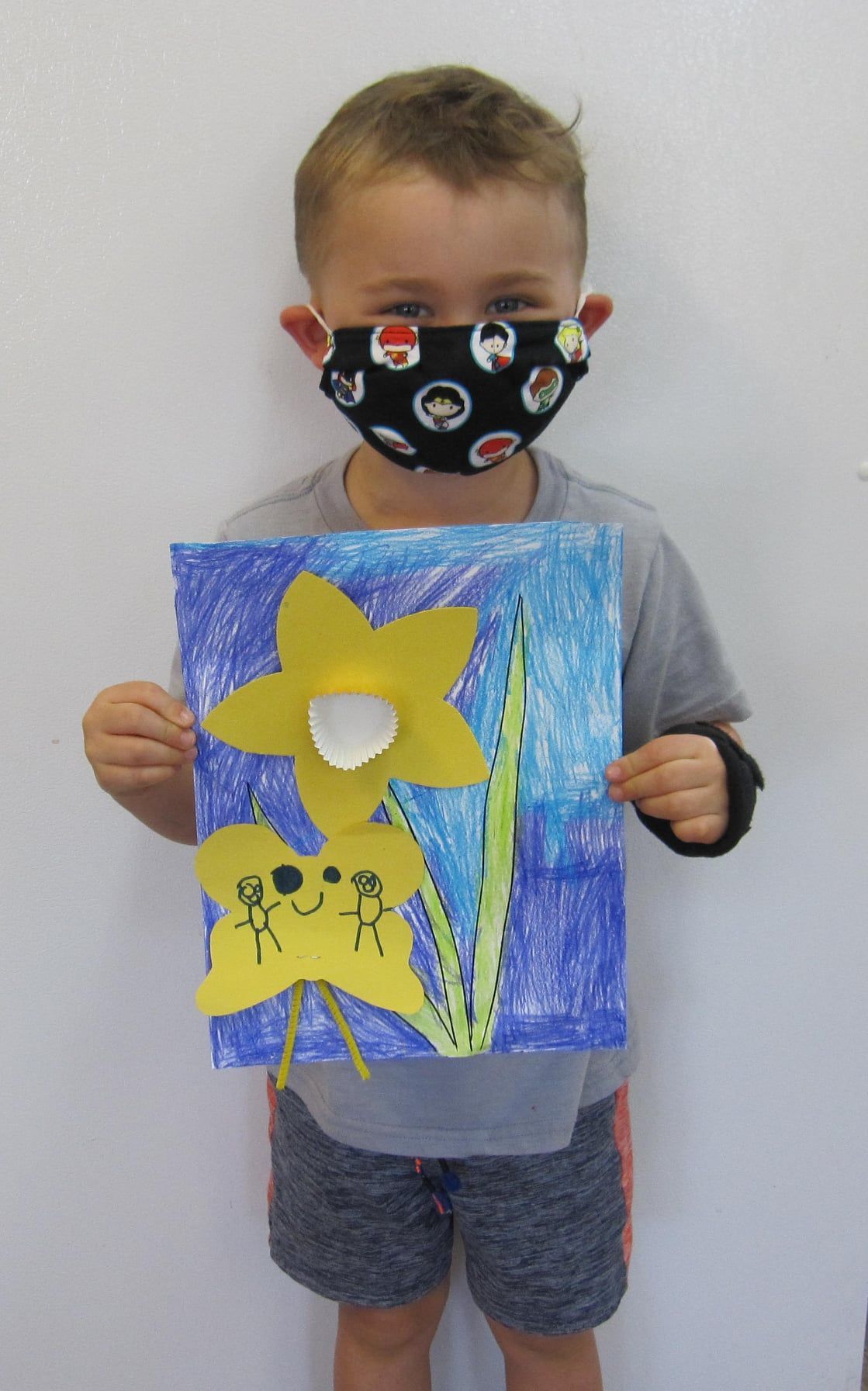 A young child wearing a mask holds a colorful daffodil drawing