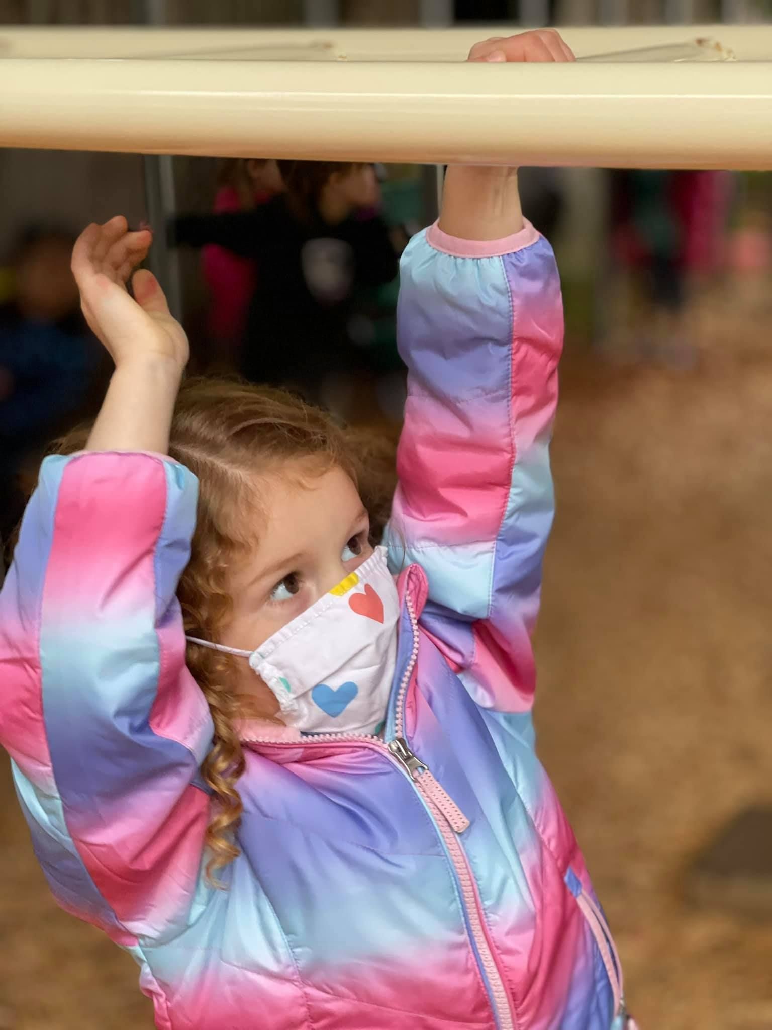 Girl in colorful jacket, mask on, hangs from monkey bars at a playground