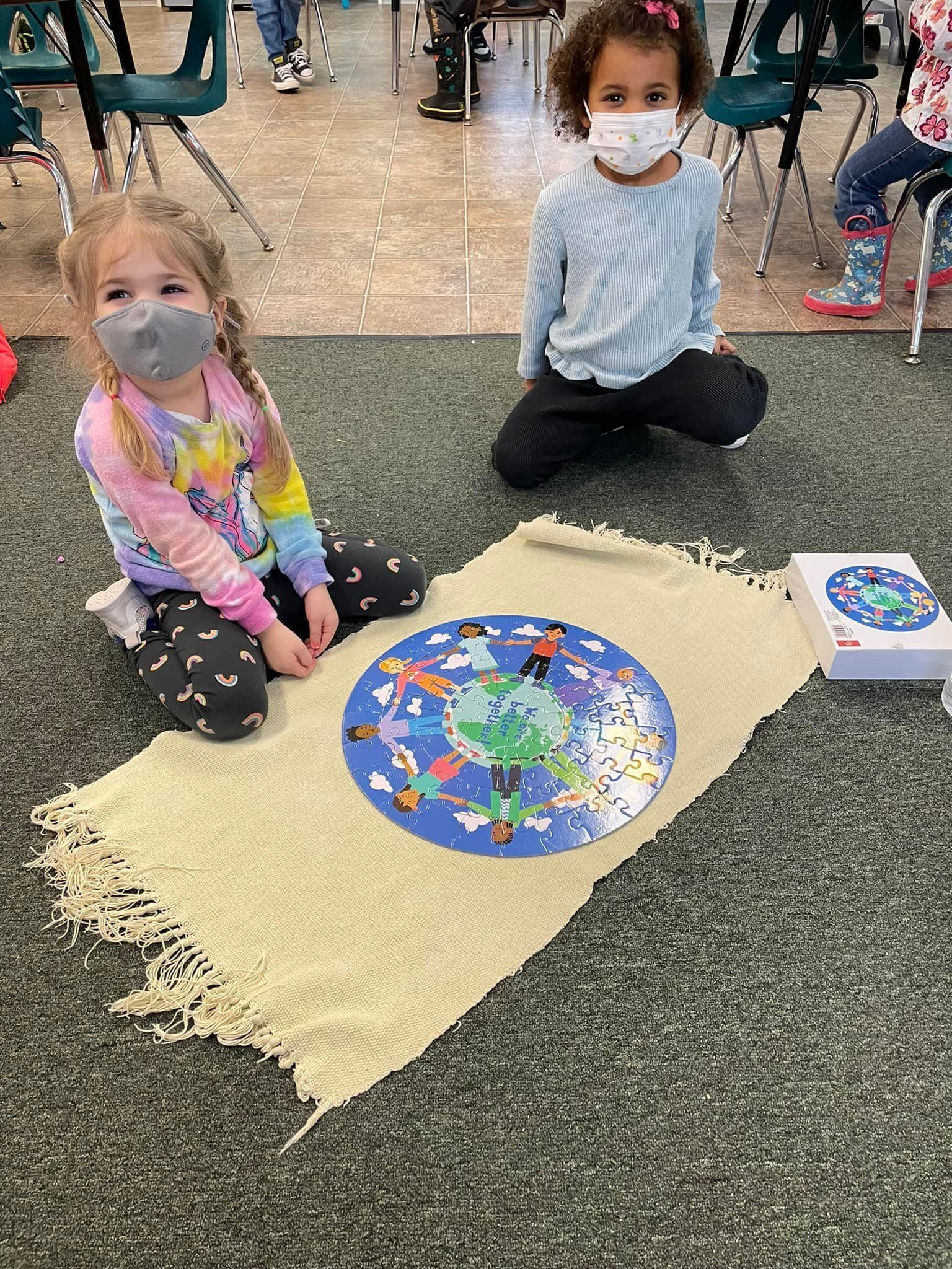 Two young girls wearing masks are working on a puzzle on a rug indoors