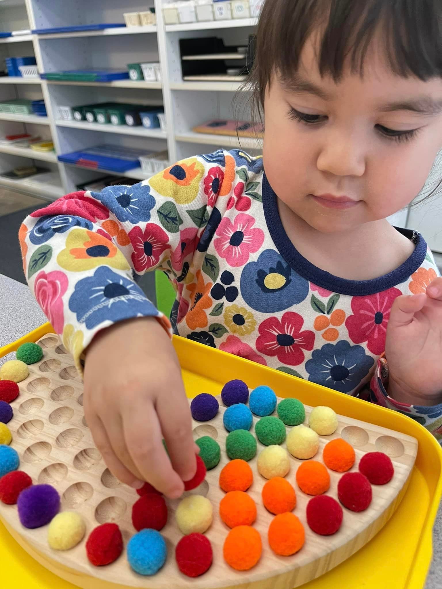 A young child in a colorful shirt sorting pom-poms by color on a wooden board