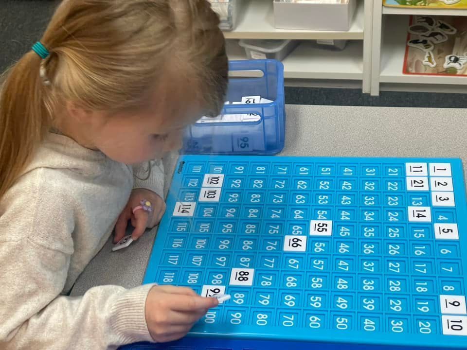 Young girl placing number tiles on a blue 100s chart in a classroom