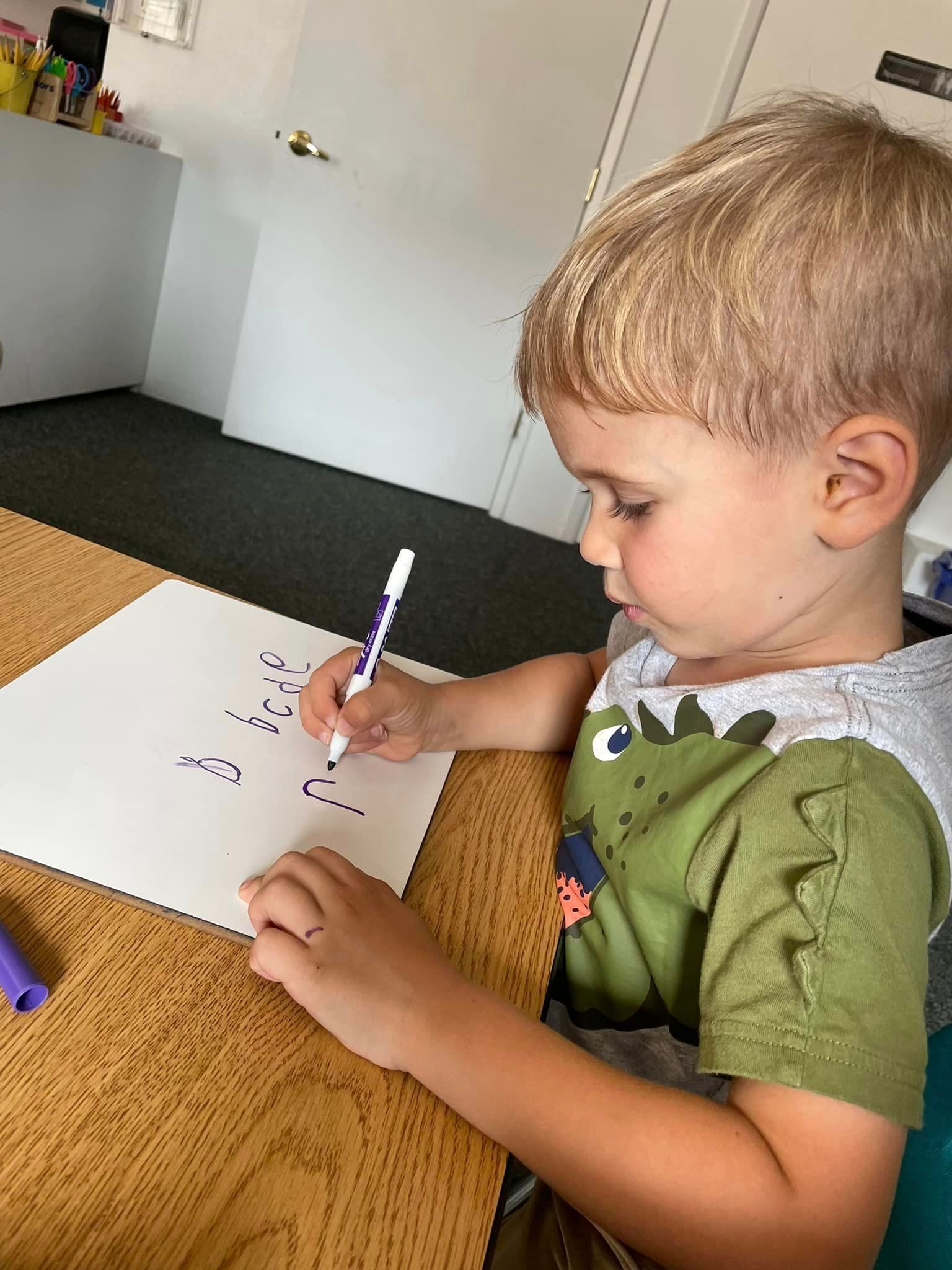 A young boy with blond hair writes with a purple marker on white paper at a table