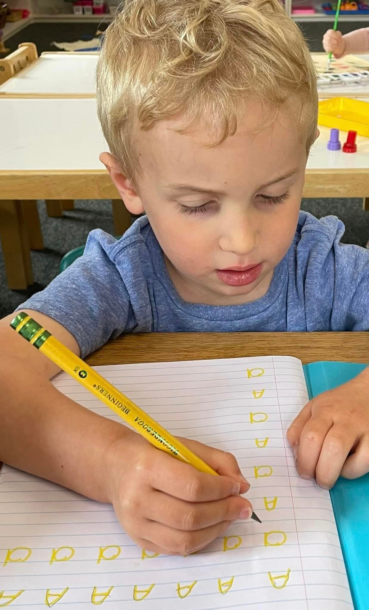 Young boy with blond hair writing on paper with a yellow pencil in a classroom setting.