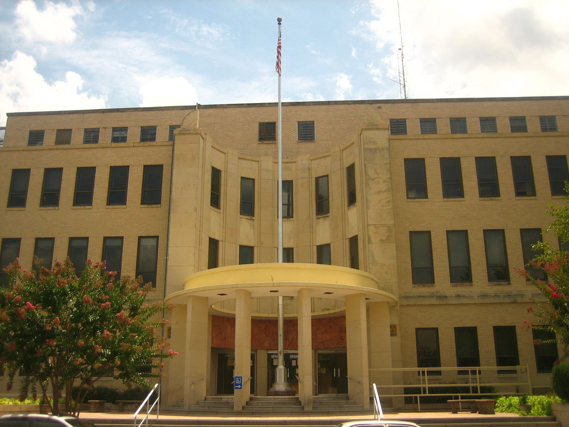 Tan beige government building with a central entrance and flagpoles, framed by trees and blue sky