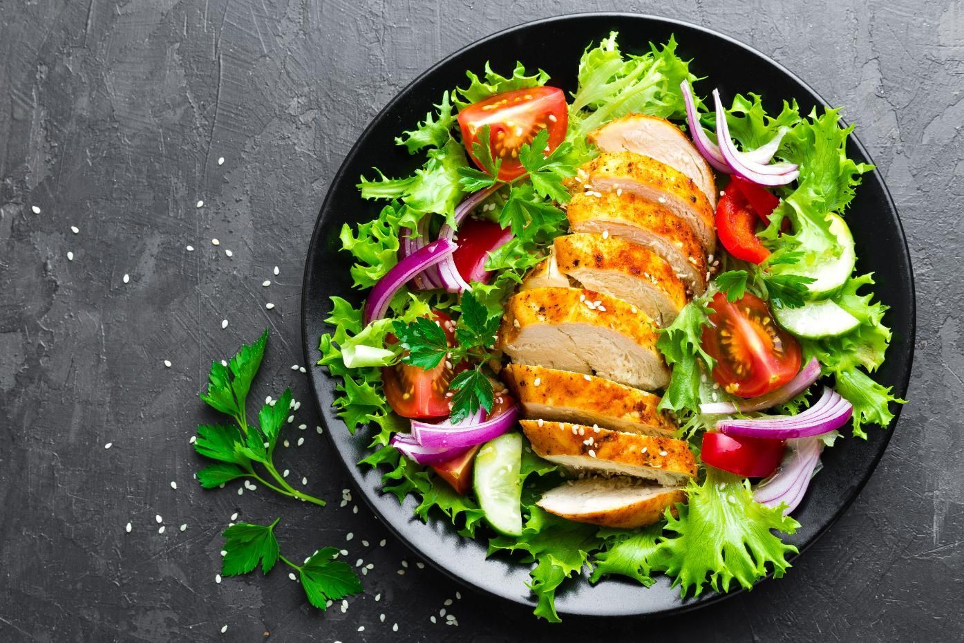 Sliced roasted chicken on leafy salad with tomatoes and red onion on a black plate