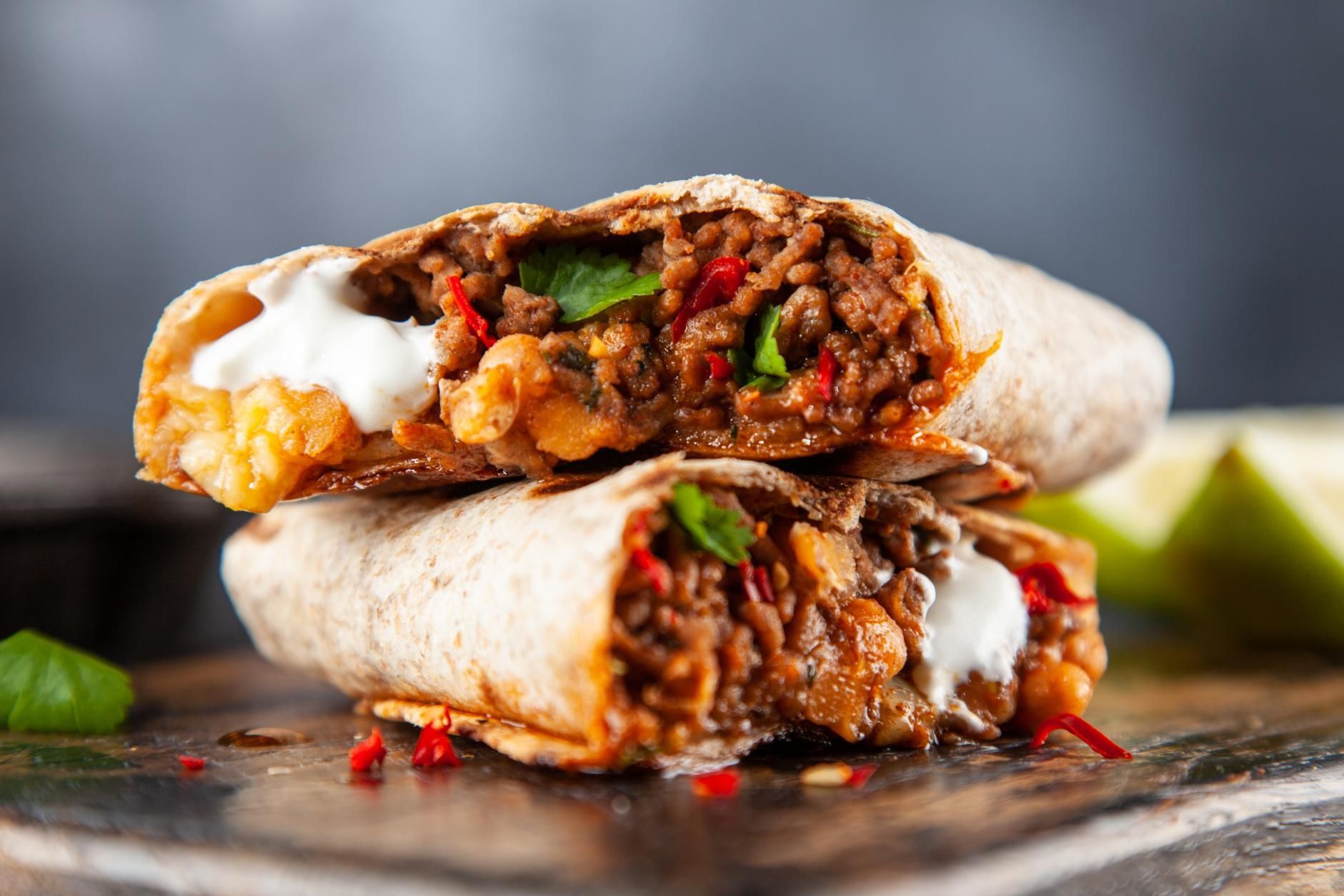 Close-up of a beef burrito cut in half, showing rice, beans, peppers, and sour cream on a dark plate.