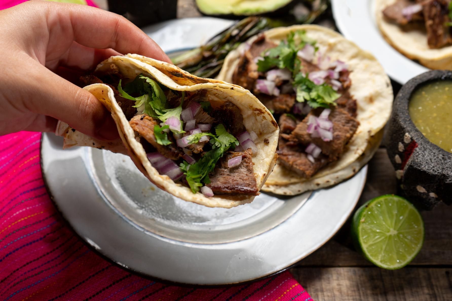 Hand holding a taco over a plate with two tacos, lime, and salsa on a table