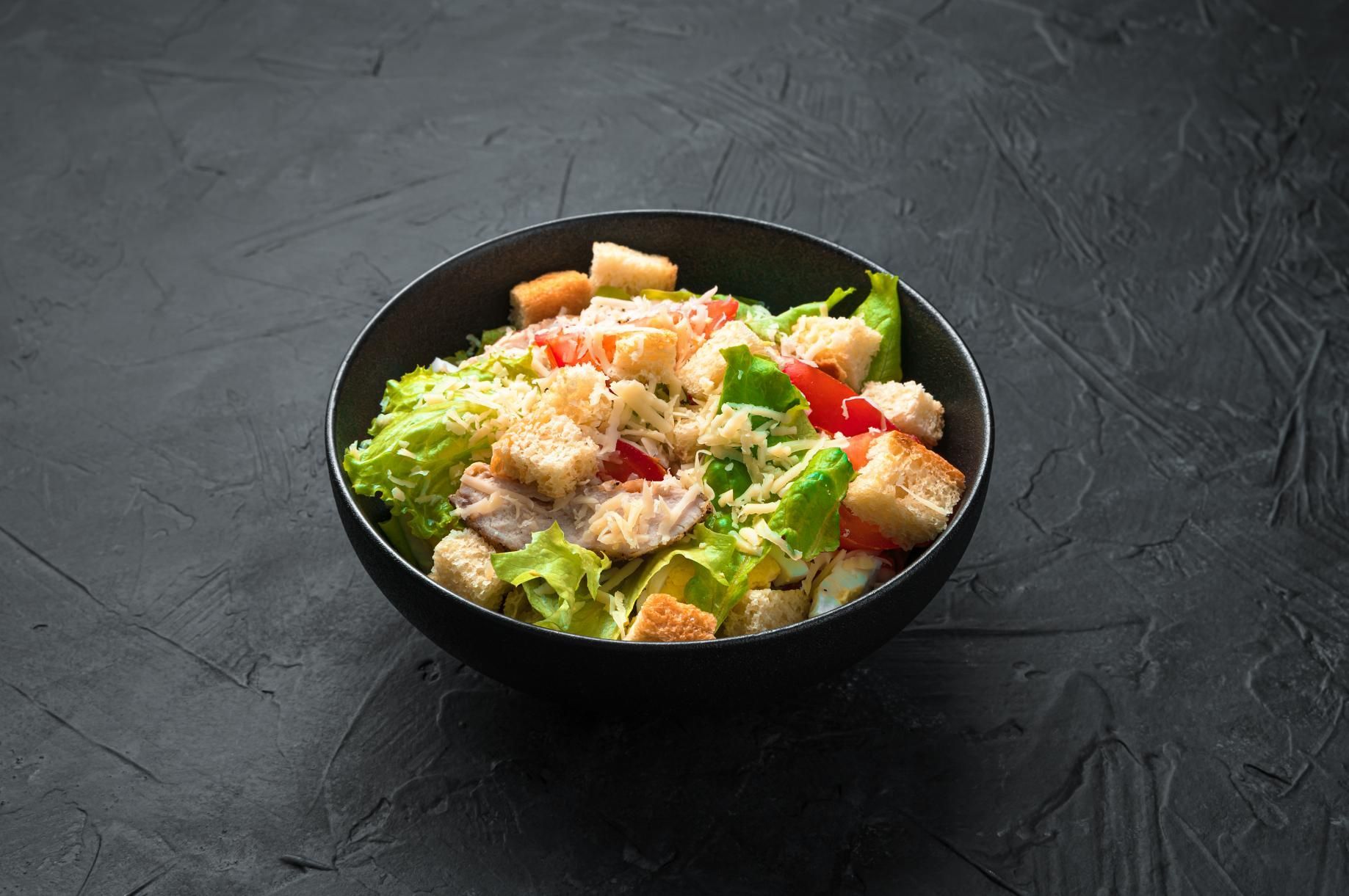 Bowl of Caesar salad with croutons, lettuce, and red peppers on a dark textured surface
