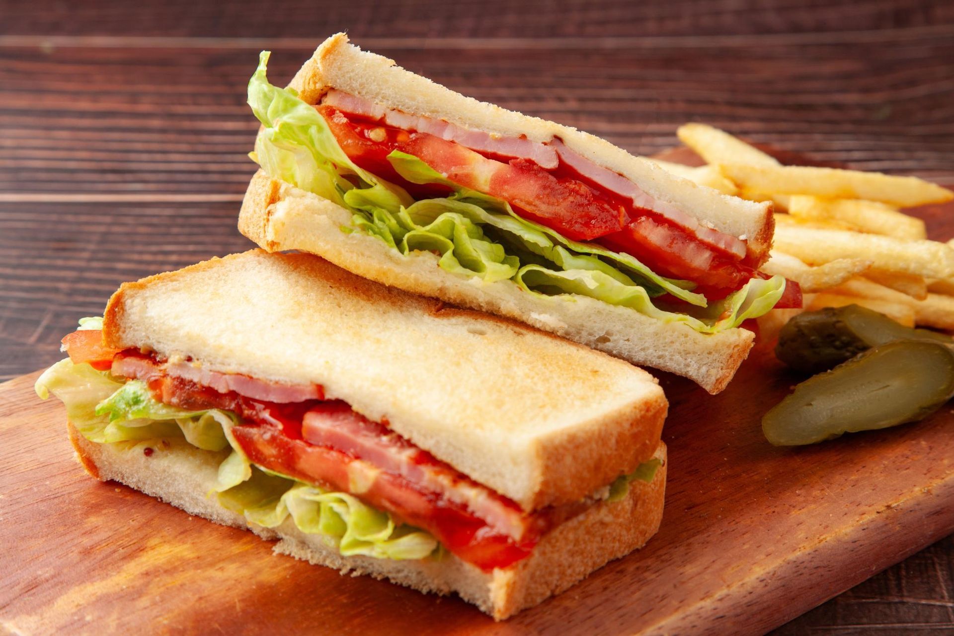 Two stacked deli sandwiches with lettuce, tomato, and pickles on a wooden board