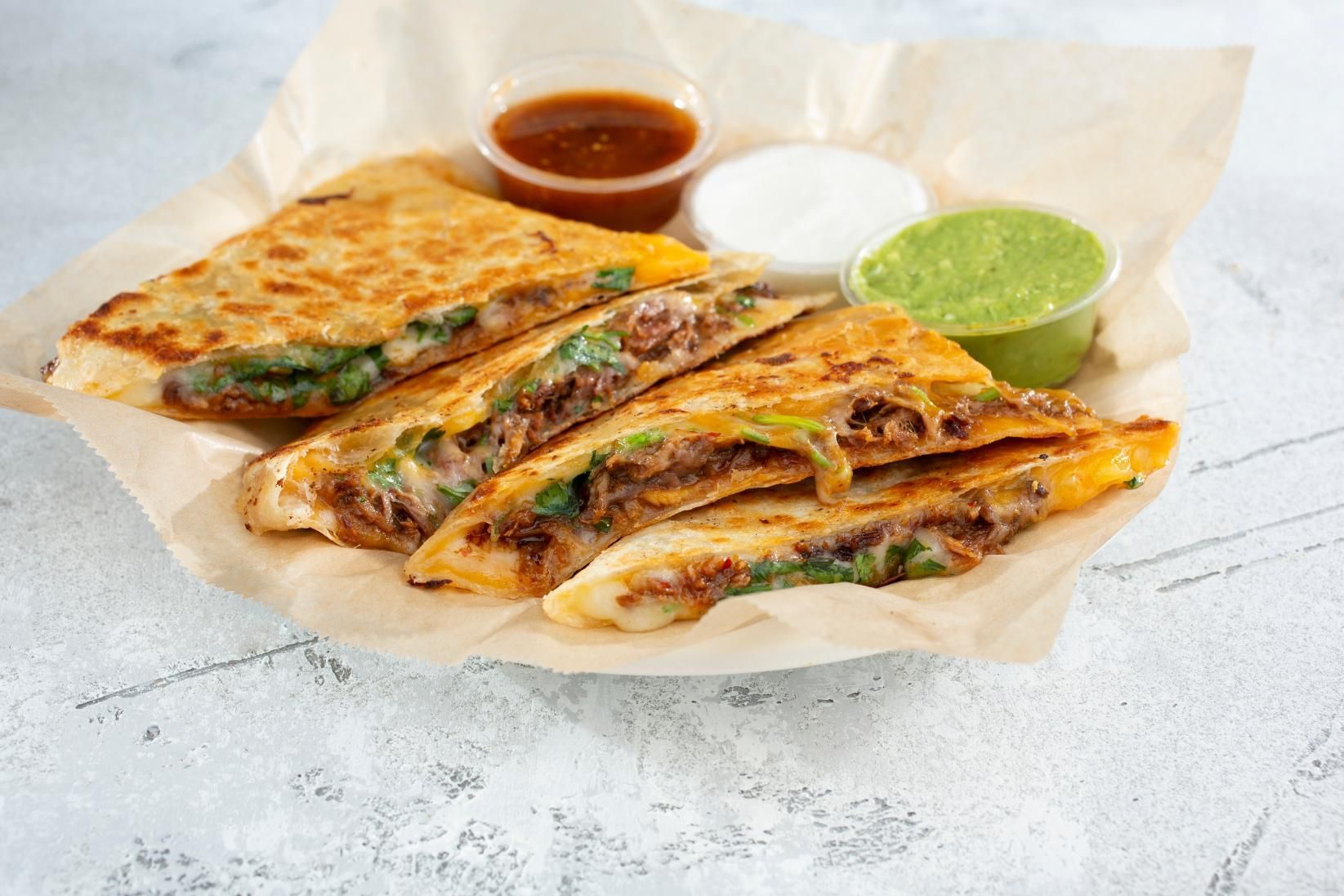 Quesadilla slices on parchment with red salsa, white sour cream, and green sauce on a light table