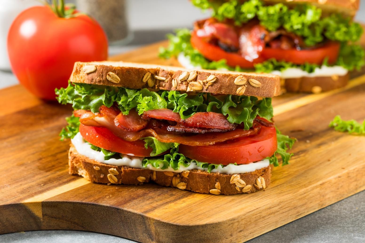 Two stacked bacon, lettuce, and tomato sandwiches on a wooden cutting board with a tomato nearby