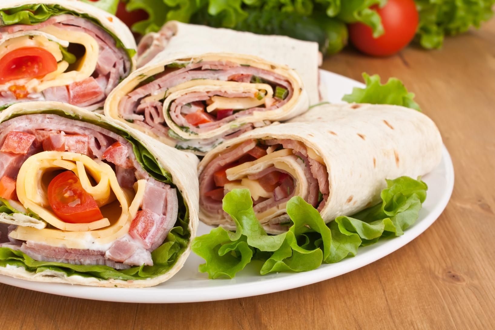 Sliced tortilla wraps with ham, cheese, and lettuce on a white plate with tomatoes and greens