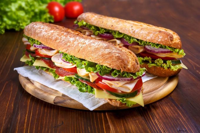 Two stacked sandwiches with lettuce, tomato, and meat on a wooden board with tomatoes in the background