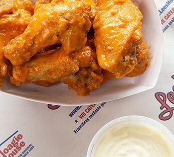 Orange buffalo wings in a white takeout tray with a small cup of ranch dipping sauce
