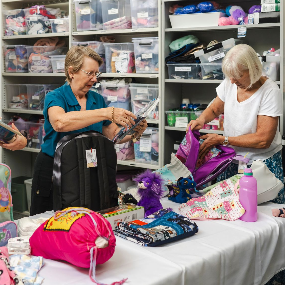 Three women sort colorful bags and purses around a table in a bright storage room.