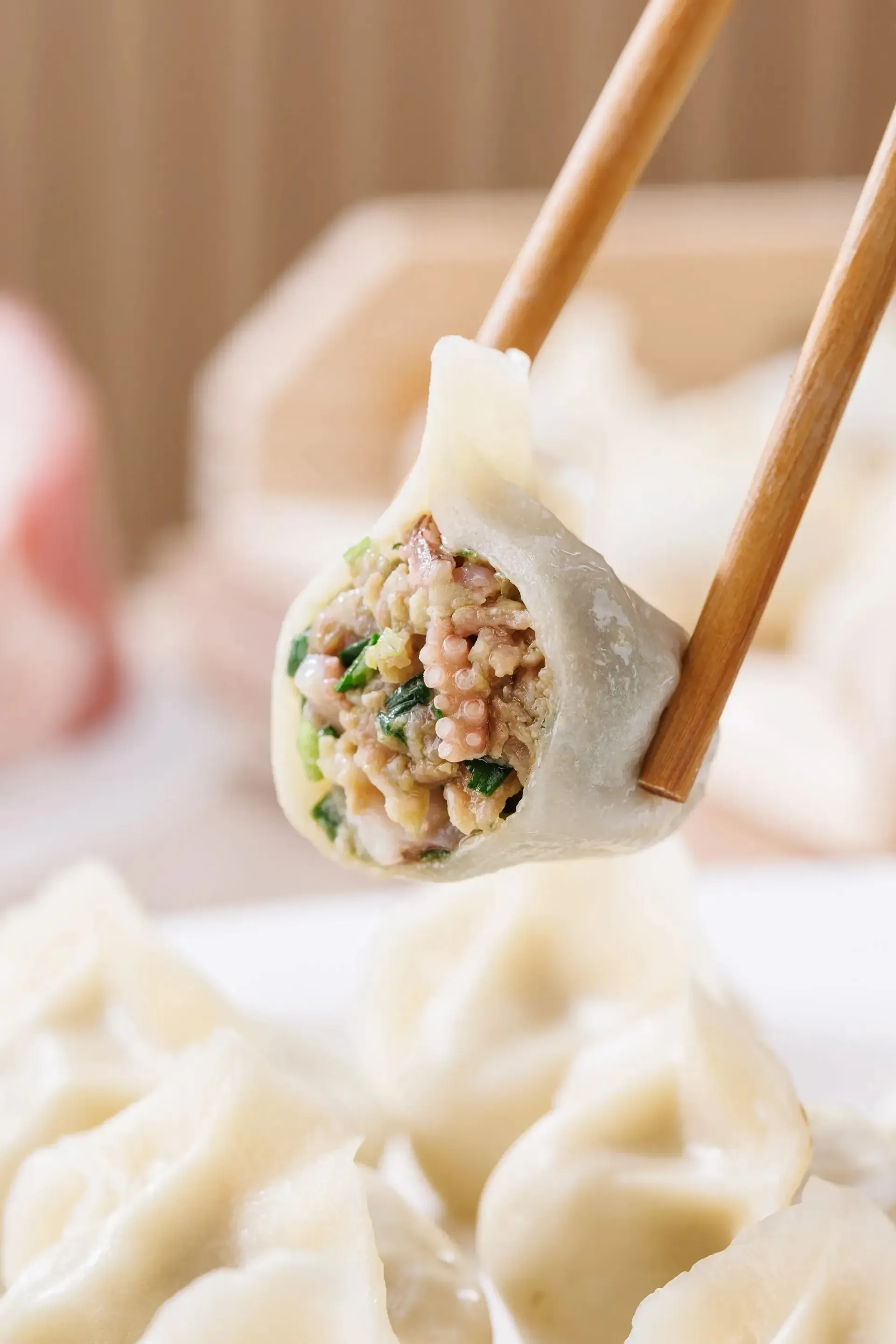 Chopsticks holding a dumpling with meat filling, with more dumplings blurred in the background