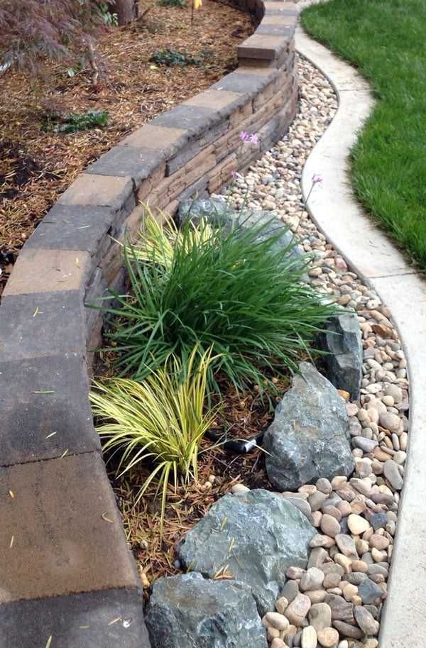 Curved garden border with stacked stone wall, pebbles, rocks, and ornamental grasses beside green lawn