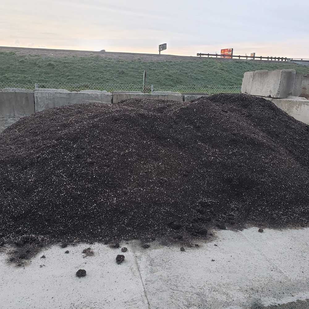 Large pile of black gravel on a concrete roadside beside a grassy embankment and fence