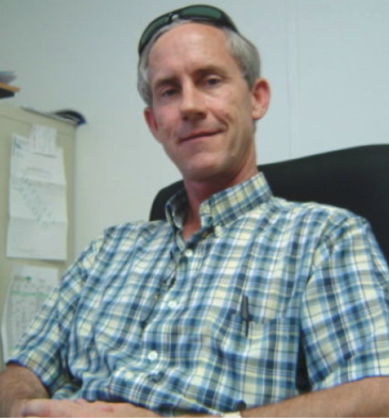 Man sitting in an office chair, wearing a blue plaid shirt and looking at the camera.
