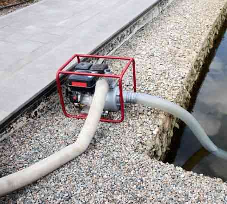 Gas-powered pump in a red frame pumping water through hoses beside a gravel canal.