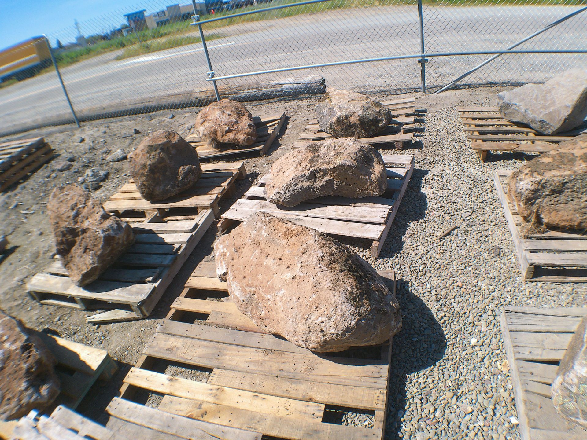 Large rocks resting on wooden pallets in a gravel yard by a chain-link fence.