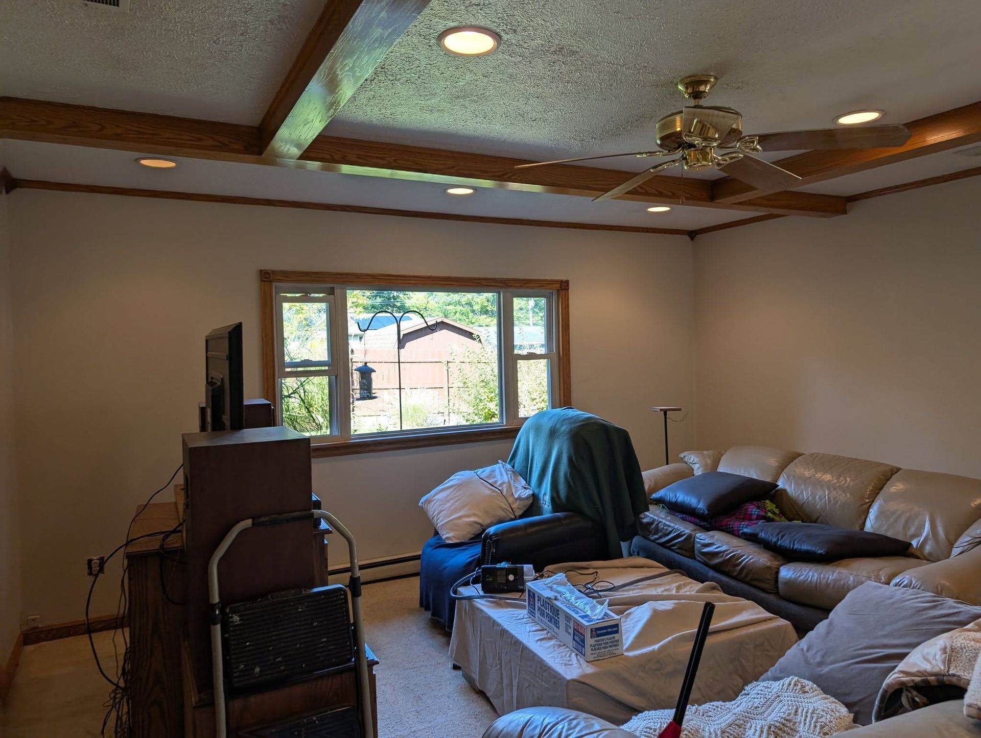 A living room with wooden ceiling beams, a large window, a sofa, a recliner with a blanket, and a television on a stand.