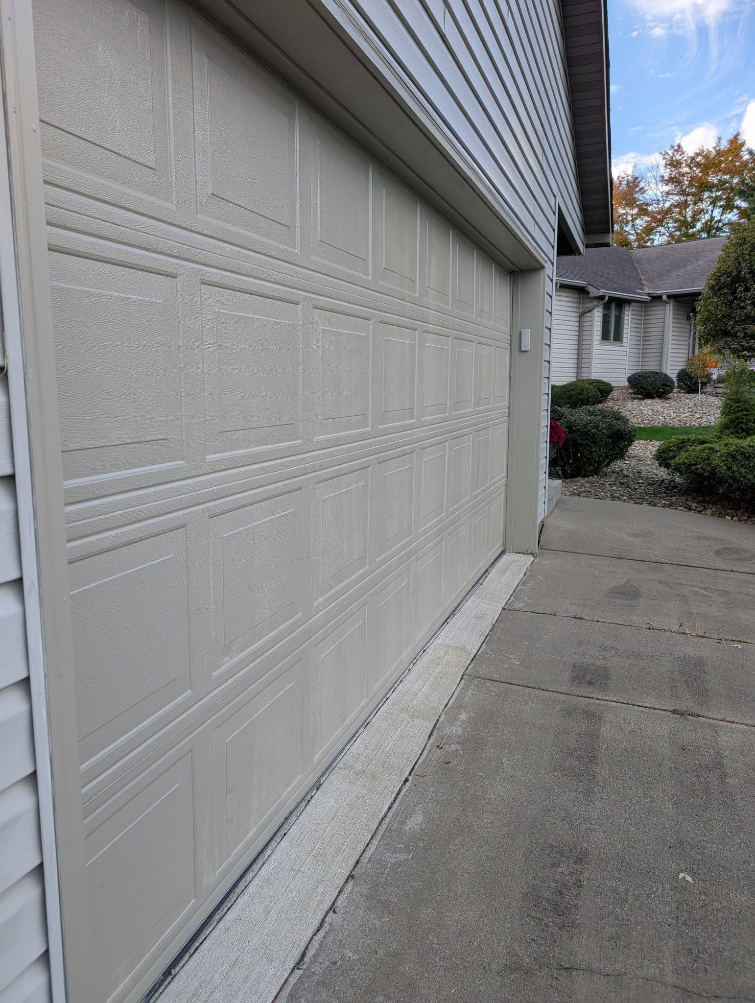 A beige, multi-panel garage door is closed against a concrete driveway with a house and bushes in the background.