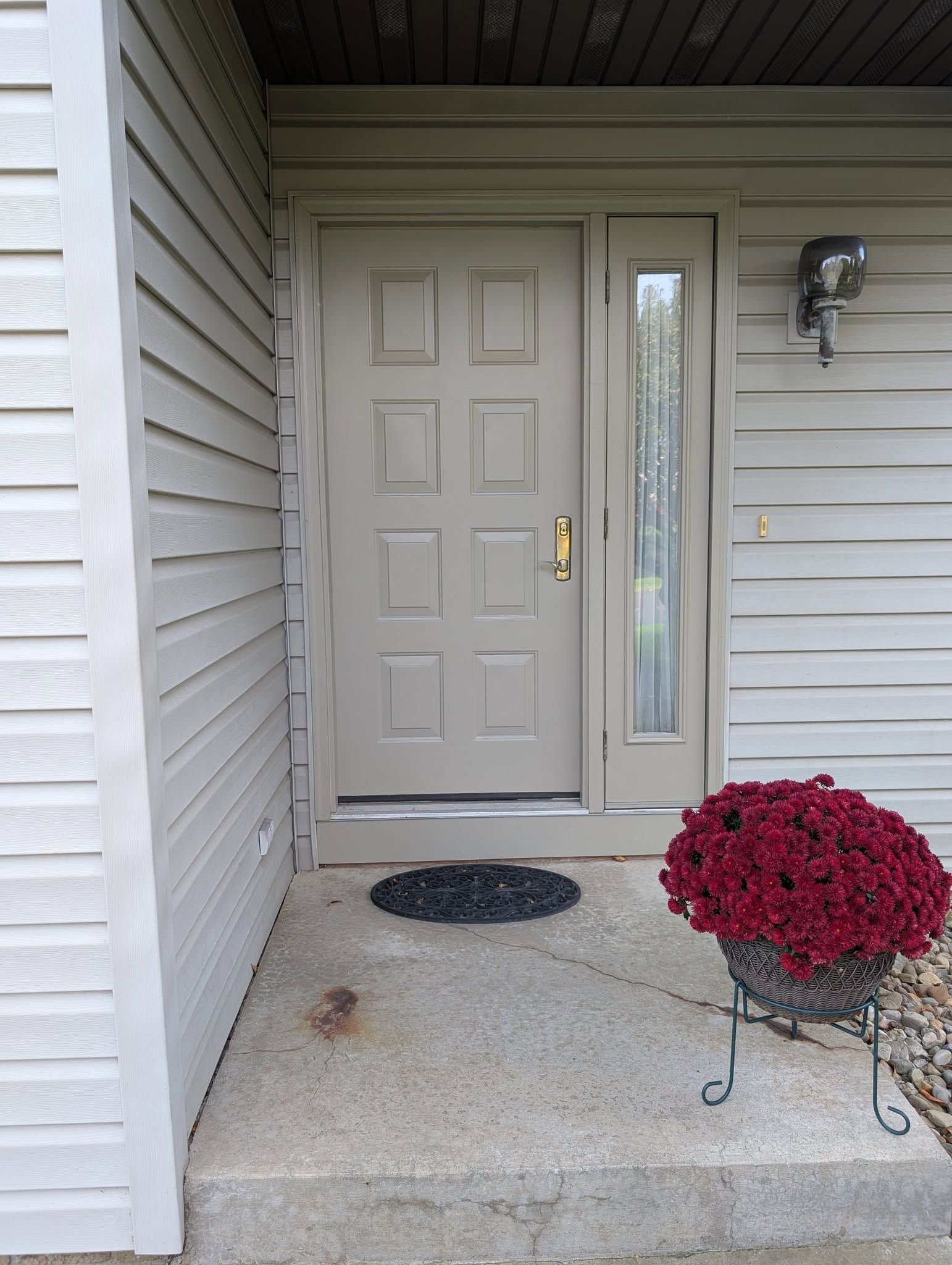 A beige front door with a side window, a dark door mat, and a potted red plant on a metal stand on a concrete porch.